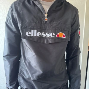 Svart vindjacka från Ellesse - Svart vindjacka från Ellesse med stor logotyp på bröstet och broderad patch på ärmen. Jackan har huva, halv dragkedja och lång ärm. Materialet är syntet och jackan har en loose passform, perfekt för streetwear. Snygg detalj med Ellesse-zipper framtill. Produkten finns på flera sidor.
