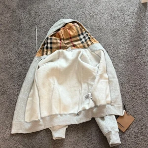 Grå Burberry hoodie med dragkedja - Snygg grå hoodie från Burberry med klassiskt rutigt foder i huvan. Tröjan har dragkedja framtill, snörning vid huvan och två fickor. Perfekt för dig som vill ha en stilren look med lyxig touch och ikoniskt mönster.