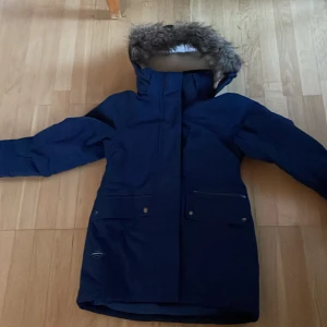 Blå vinterjacka från Didriksons - Snygg marinblå parkasjacka från Didriksons med huva och fuskpäls, perfekt för kalla dagar. Jackan har långa ärmar, flera fickor med knappar och dragkedja framtill. Insidan är fodrad med mjuk teddy och jackan har en stilren look som funkar till det mesta.