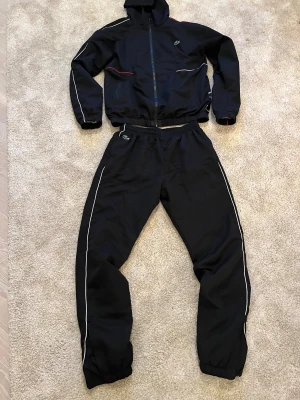 Lacoste tracksuit - Svarta trakcsuit från Lacoste med vita detaljer längs benen och elastisk midja. Materialet är lätt och syntetiskt, perfekt för sport eller chill. Klassisk Lacoste-logga på höften och snygga kontrastlinjer för en modern look. Säljer för har bytt stil. Size L men den sitter som M