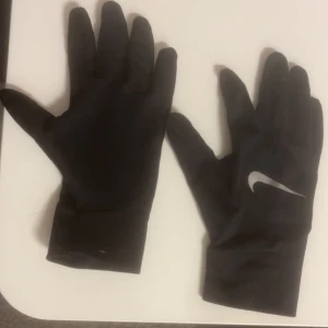 Svarta Nike handskar - Svarta handskar från Nike med klassisk vit swoosh-logga på ovansidan. Handskarna har en smidig och sportig design med lång mudd och är perfekta för träning utomhus. Materialet känns lätt och stretchigt för bra passform.