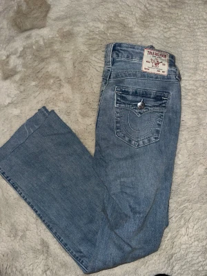 True Religion Becca Bootcut Jeans - Snygga ljusblå jeans från True Religion, modell Becca Mid Rise Bootcut. Klassisk bootcut-passform med coola detaljer som silverfärgade knappar och broderade bakfickor. Jeansen är tillverkade i mjukt denim. Endast använda 1 gång.