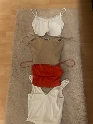 Fyra trendiga croppade toppar - Säljer fyra snygga croppade toppar: en vit spaghettitopp med spetskant, en ribbad beige linnetopp, en röd axelbandslös topp med tunna band och en vit v-ringad topp. Perfekta för sommaren och att mixa och matcha till olika outfits.