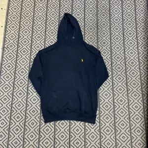 Marinblå hoodie från Polo Ralph Lauren - Snygg marinblå hoodie från Polo Ralph Lauren med klassisk gul broderad logga på bröstet. Tröjan har huva med metallöljetter och en stor magficka framtill. Perfekt för dig som gillar stilren och enkel design med premiumkänsla.axelbredd: 51 cm och längd: 63 cm – motsvarar tröjan ungefär en herrstorlek M (Medium) i de flesta märken, möjligen S/M beroende på passform (t.ex. om den är slim fit eller regular fit).skickar samma dag!