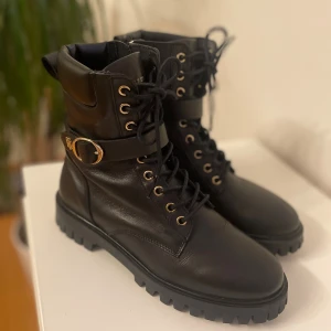 Svarta boots från Tommy Hilfiger - Säljer ett par svarta boots från Tommy Hilfiger i skinn med grov platt sula och snörning. De har guldfärgade detaljer, spänne med logotyp på sidan och rund tå. Nyskick 