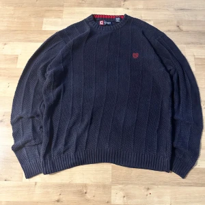 Mörkblå stickad tröja från Chaps - Säljer en mörkblå stickad tröja från Chaps med diskret röd broderad logga på bröstet. Tröjan har ett grovt, randigt stickat mönster och ribbade muddar vid ärmslut och nederkant. Perfekt för dig som gillar klassisk och avslappnad stil.