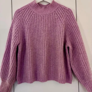 Ljusrosa stickad tröja med ribbade muddar - Supermysig ljusrosa stickad tröja med grov struktur och ribbade muddar vid ärmslut och hals. Tröjan har en relaxed passform och är perfekt för kyliga dagar. Enkel att styla med jeans eller kjol för en chill look.