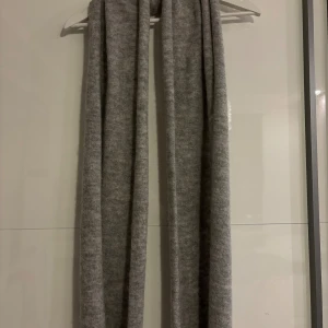 Lång grå stickad halsduk - Säljer en supermjuk och lång grå halsduk i stickad kvalitet. Perfekt att svepa runt halsen för extra värme under kyliga dagar. Enkel och stilren design som passar till de flesta jackor och outfits.
