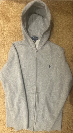  Polo Ralph Lauren zip up - Snygg grå hoodie från Polo Ralph Lauren med dragkedja och klassisk blå logga på bröstet. Tröjan har huva, långa ärmar och är tillverkad i mjuk bomull med inslag av polyester. Perfekt för chill dagar och enkel att matcha med jeans eller joggers.