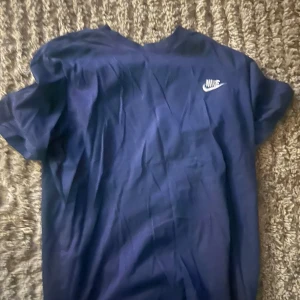 Blå Nike t-shirt med logga - Snygg blå t-shirt från Nike med klassisk vit logga på bröstet. Modellen är enkel med rund hals och korta ärmar, perfekt för en chill och sportig stil. Materialet känns mjukt och skönt mot huden.