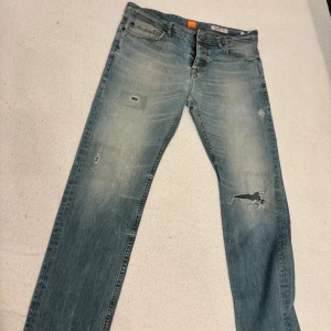 Blå jeans från Hugo Boss, storlek 33/32 - Snygga ljusblå jeans från Hugo Boss med slitna detaljer och hål på benen för en cool, avslappnad look. Klassisk femficksmodell med orange Boss-märke i midjan och kontrastsömmar. Passar dig som gillar streetstyle och vill sticka ut.