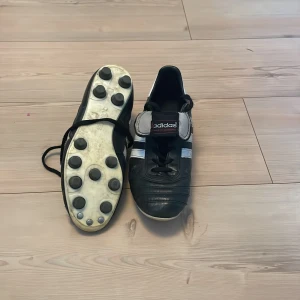 Copa mundial  - Klassiska svarta fotbollsskor från Adidas med vita detaljer och snörning. Skorna har rund tå och platt sula med dobbar för extra grepp på planen. Märket syns tydligt på tungan och de är tillverkade i skinn för bra komfort och hållbarhet.