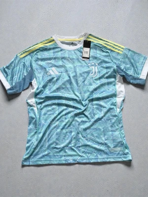 Juventus Adidas Third Jersey - Authentic Juventus football jersey by Adidas, featuring a striking teal/blue graphic pattern with classic Adidas shoulder stripes and embroidered Juventus crest. This standout shirt has a modern-retro look that’s perfect for collectors, fans, or streetwear styling.