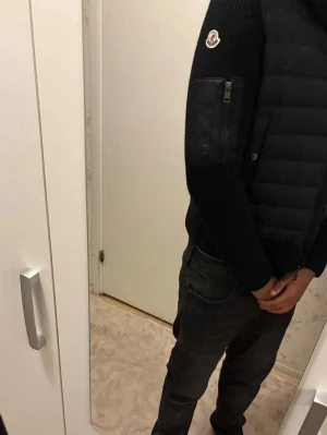 Svart Moncler pufferjacka med huva - Svart pufferjacka från Moncler med stickade ärmar och huva. Jackan har ribbade muddar, dragkedja framtill och fickor med knappar. Moncler-logga på ena ärmen och en extra ficka med dragkedja. Perfekt för dig som vill ha en snygg och varm jacka med streetkänsla.