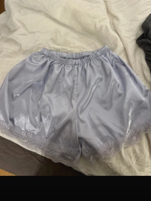 Ljuslila silkesliknande shorts med spets - Säljer ett par ljuslila shorts i silkesliknande material med elastisk midja och fina spetsdetaljer längst ner. Perfekta för en chill look eller som sovplagg. Supersköna och mjuka mot huden.