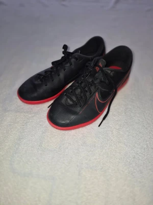 Nike svart/röda innefotbollsskor strl 39 - Säljer ett par svarta Nike innefotbollsskor med röda detaljer och sula. Skorna har snörning, låg profil och är tillverkade i syntetmaterial. Swoosh-loggan syns tydligt på sidan och sulan har greppvänligt mönster. Perfekta för inomhusspel.