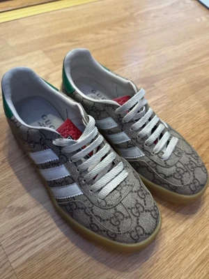 Gucci x Adidas Gazelle sneakers beige - Säljer ett par Gucci x Adidas Gazelle sneakers i beige med GG-monogram över hela skon, vita ränder på sidorna och grön häl med gul logga. Skorna har vit snörning, röd detalj på plösen och gummisula. Riktigt snygga och unika sneakers för dig som vill sticka ut.