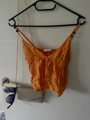 Orange glittrig linnetopp Pull&Bear - Säljer en orange linnetopp från Pull&Bear med tunna axelband och v-ringning. Toppen har glittriga detaljer i tyget och en lätt rynkad struktur. Perfekt för sommarens alla tillfällen och ger en fräsch vibe till din outfit.