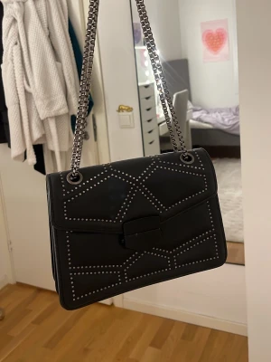 Svart axelväska med nitar - Snygg svart axelväska i skinnimitation med coola silverfärgade nitar i geometriskt mönster. Kedjerem i metall och stilren fyrkantig form. Perfekt storlek för det viktigaste och stängs med ett lock och spänne framtill.