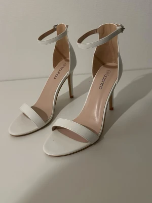 Vita klackade sandaler från Boohoo - Säljer ett par stilrena vita sandaler från Boohoo med smal rem över tårna och justerbart ankelband med spänne. Skorna har hög klack och öppen tå, perfekta för att lyfta din outfit. Tillverkade i syntetmaterial med en elegant och minimalistisk design.