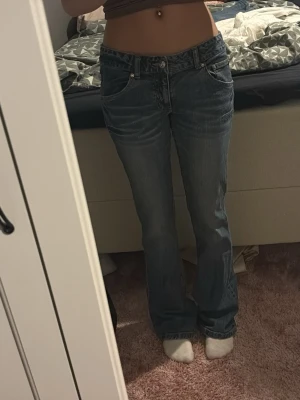 Blå bootcut jeans med krona - Säljer ett par blå bootcut jeans med låg midja och snygga broderade kronmotiv på bakfickorna. Jeansen har klassisk femficksdesign och coola detaljer med nitar runt brodyren. Perfekta för dig som vill sticka ut lite extra.