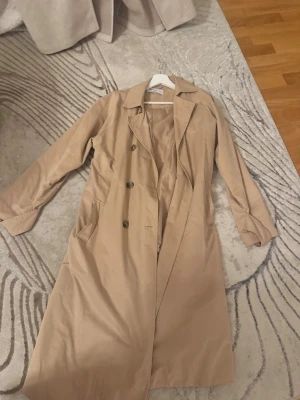 Beige trenchcoat från Ellos - Stilren beige trenchcoat från Ellos med klassisk krage, knappar framtill och bälte i midjan. Jackan har långa ärmar och fickor på sidorna. Perfekt för dig som vill ha en tidlös och snygg look till vardags.