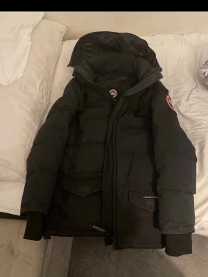 Svart dunjacka från Canada Goose M - Säljer en svart dunjacka från Canada Goose med huva och dragkedja. Jackan har två stora fickor framtill, ribbade muddar och klassisk Canada Goose-logga på ärmen. Perfekt för kalla dagar och riktigt snygg streetstil.