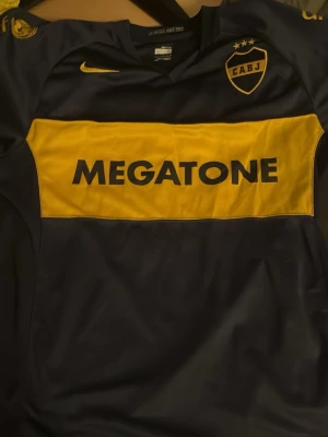 Boca Juniors fotbollströja Nike - Säljer en klassisk Boca Juniors fotbollströja från Nike med gul tvärrand och trycket MEGATONE på bröstet. Tröjan är marinblå med gula detaljer och klubbmärke på bröstet. Perfekt för dig som älskar fotboll och vill sticka ut på planen. 