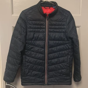 Mörk blå pufferjacka från Jack & Jones - Säljer en svart pufferjacka från Jack & Jones med röd insida och snyggt märke på ärmen. Jackan har quiltad design, hel dragkedja framtill och står upp lite i kragen. Perfekt för kyliga dagar och riktigt skön att ha på sig.Har ett jätte litet hål på ena armen.