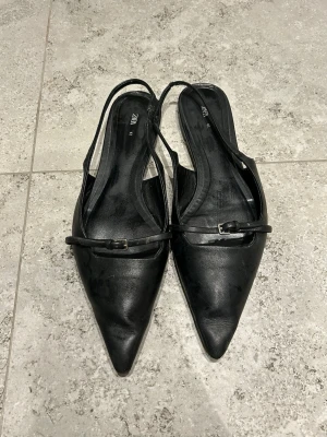 Svarta ballerinaskor från Zara - Säljer ett par svarta ballerinaskor från Zara i storlek 41. Skorna har spetsig tå, smal rem med spänne över foten och är tillverkade i skinn. Snygg minimalistisk design som passar till många olika stilar.