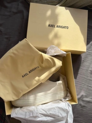 Vita sneakers från Axel Arigato - Säljer ett par stilrena vita sneakers från Axel Arigato i skinn. Skorna har minimalistisk design utan snörning, med diskret logga i guld på sidan och tjock vit sula. Perfekta för dig som gillar cleana och moderna vibes.