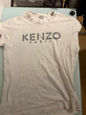 Vit Kenzo t-shirt med logga - Vit t-shirt från Kenzo med stor svart logga på bröstet. Klassisk rund halsringning och korta ärmar. Tillverkad i mjuk bomull som känns skön mot huden. Perfekt för dig som gillar stilrena och trendiga plagg med designer-touch.
