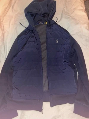 Mörkblå pufferjacka från Polo Ralph Lauren - Säljer en mörkblå pufferjacka från Polo Ralph Lauren med huva och dragkedja. Jackan har quiltade partier framtill, långärmad design och den klassiska loggan broderad på bröstet. Perfekt för kyliga dagar och riktigt snygg till jeans eller sweatpants.