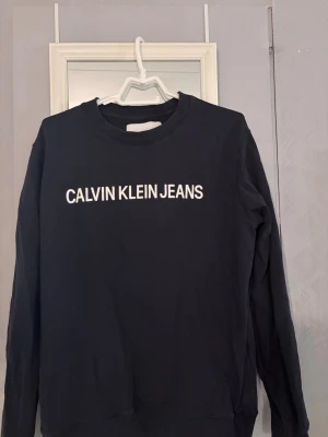 Svart sweatshirt från Calvin Klein Jeans - Säljer en svart sweatshirt från Calvin Klein Jeans i storlek XL. Tröjan har klassisk rund hals, långa ärmar och vit logga tryckt framtill. Tillverkad i mjuk bomull 