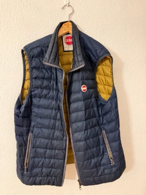 Mörkblå dunväst från Colmar - Säljer en mörkblå quiltad dunväst från Colmar med gul insida. Västen har två sidofickor med dragkedja och en hög krage. Klassisk Colmar-logga på bröstet och nedtill på ryggen. Perfekt för lager-på-lager och chill stil.