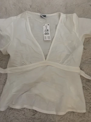 Vit omlottopp från Gina Tricot - Säljer en vit omlottopp från Gina Tricot i storlek S. Toppen har korta ärmar, djup v-ringning och knytband i midjan som ger en snygg siluett. Perfekt till jeans eller kjol för en fräsch look.