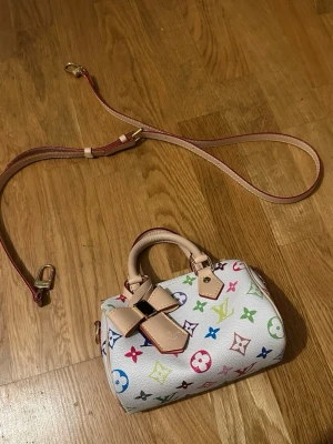 Louis Vuitton Murakami speedy nano handväska - Säljer en ikonisk Louis Vuitton Murakami handväska i vitt med färgglatt monogrammönster. Väskan har ljusa skinnhandtag, avtagbar axelrem och en snygg rosettdetalj framtill. Perfekt storlek för det viktigaste och riktigt unik design.