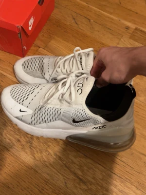 Nike Air Max 270 vita sneakers - Nike Air Max 270 sneakers i vitt med svarta detaljer och stor synlig Air-enhet i hälen. Ovandelen är i mesh med ventilerande mönster och snörning framtill. Snygg och sportig look som passar perfekt till streetwear och träning.