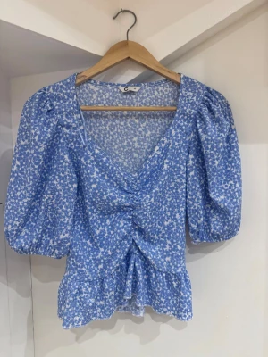 Blommig blå blus med puffärm XS - Säljer en söt blommig blus från C i storlek XS. Blusen är blå med vita blommor, har puffiga korta ärmar, v-ringning och volang nedtill. Materialet känns lätt och luftigt, perfekt för vår och sommar. Supersnygg till jeans eller kjol!