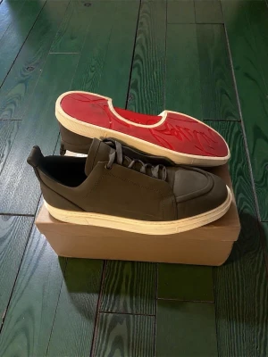 Christian Louboutin sneakers med röd sula - Säljer ett par snygga sneakers från Christian Louboutin i mörkbrunt skinn med vita sulor och den ikoniska röda undersidan. Skorna har snörning och en stilren, chunky design som sticker ut. Perfekta för dig som vill ha något exklusivt och trendigt.
