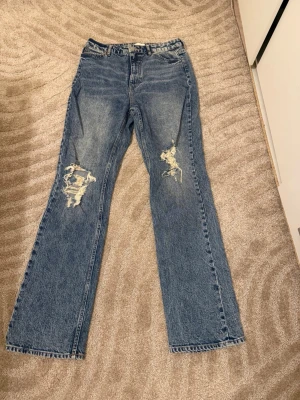 Blå raka jeans med slitningar - Säljer ett par blå jeans med raka ben och slitna detaljer på båda knäna. Klassisk femficksmodell i jeansmaterial, perfekt för en avslappnad och trendig look. Passar dig som gillar vintage vibe och streetstyle.