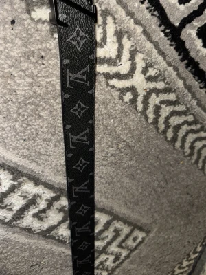 Svart monogram-bälte - Säljer ett svart bälte  med klassiskt monogrammönster och stor -logga i metall som spänne. Bältet har en lyxig känsla och är gjort i präglat skinn med mörka detaljer. Perfekt accessoar för att lyfta vilken outfit som helst.