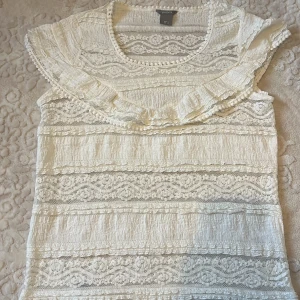 Vit spetsblus med volang från Lindex - Säljer en söt vit blus från Lindex med spetsdetaljer och volang runt axlarna. Blusen har rund halsringning och är ärmlös, med genombrutna spetsmönster i horisontella ränder. Perfekt för dig som gillar romantisk och feminin stil.