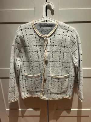 Grå rutig kofta med pärlknappar - Säljer en grå kofta med svart rutmönster och dekorativa pärlknappar. Koftan har två fickor framtill och fina flätade detaljer längs kanterna. Långärmad och perfekt för lager på lager under kyliga dagar.