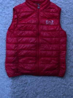 Röd dunväst från Emporio Armani EA7 - Snygg röd dunväst från Emporio Armani EA7 med quiltad design och dragkedja framtill. Västen har hög krage och logga på bröstet. Perfekt att slänga över en hoodie för en unik och snygg stil! Skriv om frågor svarar så snabbt som möjligt