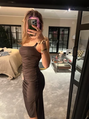Brun tight långklänning med smala axelband - Säljer en brun långklänning med smala axelband och figurnära passform. Klänningen har en enkel och stilren design med lätt rynkade detaljer längs sidorna och en liten slits nertill. Perfekt för dig som gillar en minimalistisk och elegant look.