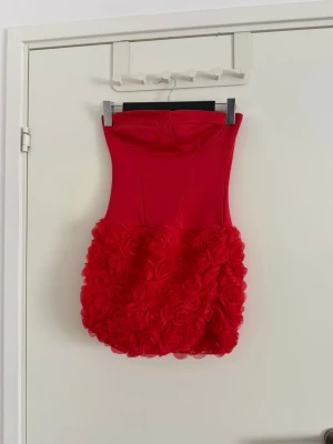 Röd axelbandslös kortklänning - Supersnygg röd axelbandslös kortklänning med tight överdel och voluminös kjol täckt av 3D-blommor. Kjolen har en unik struktur med rosdetaljer som verkligen sticker ut. Perfekt för dig som vill ha en statement piece till nästa fest. Aldrig använd då den är lite för stor för mig. Den är storlek xs men skulle säga om man vill att den sitter tajt så är de mer än S