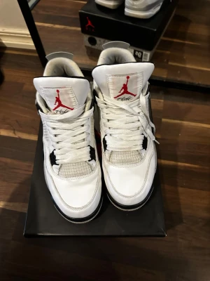 Nike Air Jordan 4 White Cement - Nike Air Jordan 4 White Cement med vit ovandel i skinn, grå och svarta detaljer samt röda inslag på loggan och yttersulan. Klassisk meshpanel på sidorna, synlig snörning och ikonisk Nike Air-logga på hälen. Snygg retrostil som alltid levererar.