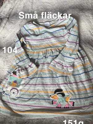 Randig barnklänning med hundmotiv - Söt klänning från Polarn O. Pyret i grått med färgglada ränder i blått, gult, lila och grönt. Klänningen har volangdetaljer vid ärmslut och roliga applikationer med hund på rullskridskor och en ficka med glassmotiv. Perfekt för lek och bus!🌸 Små fläckar. Storlek 104