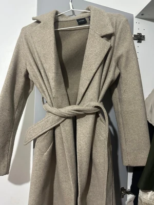 Beige kappa från Vero Moda - Säljer en stilren beige kappa från Vero Moda med klassisk krage och knytskärp i midjan. Jackan är lång och har diskreta fickor på sidorna. Perfekt för dig som vill ha en enkel men snygg look till höst och vinter.